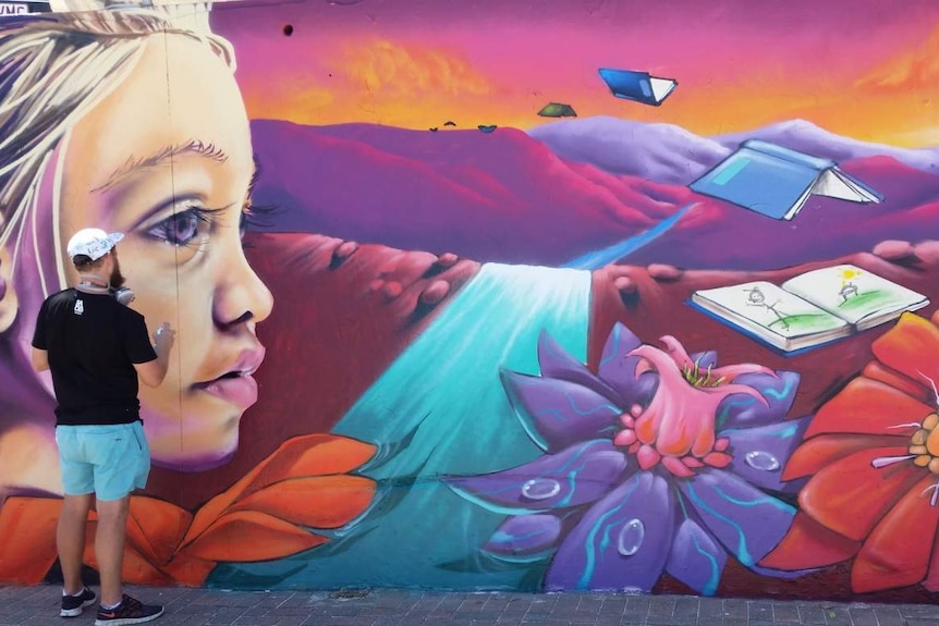 An artist works on a mural which shows a young girl looking out at flowers and books.