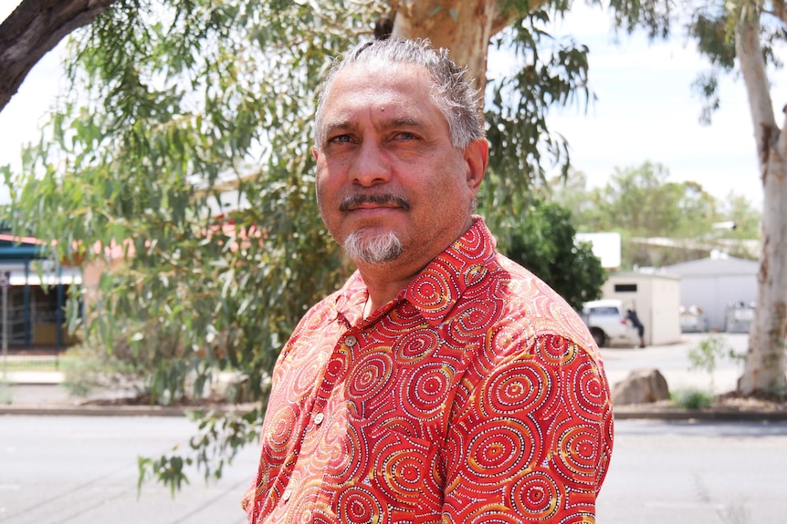 A man wearing a red shirt looks seriously at the camera. He has a goatie and his hair is pulled back off his face.