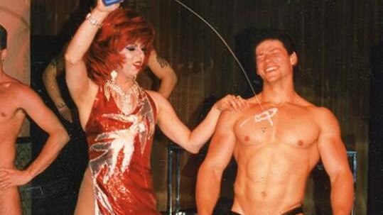 drag queen in sequined frock arm raised pouring oil on torso of male stripper in black bathers
