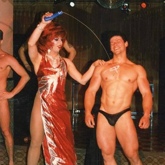 drag queen in sequined frock arm raised pouring oil on torso of male stripper in black bathers