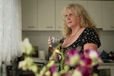 a woman with long curly blonde hair looks out a kitchen window holding a glass of water