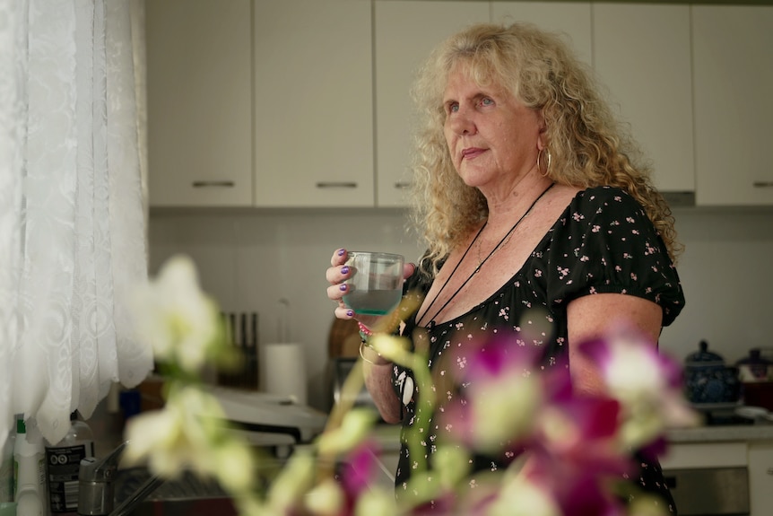 a woman with long curly blonde hair looks out a kitchen window holding a glass of water