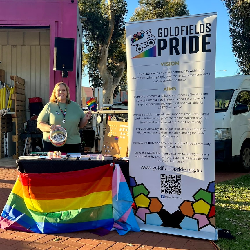 A woman n a green top holding a bucket and standing next to a Goldfields Pride banner and a desk draped in the pride flag