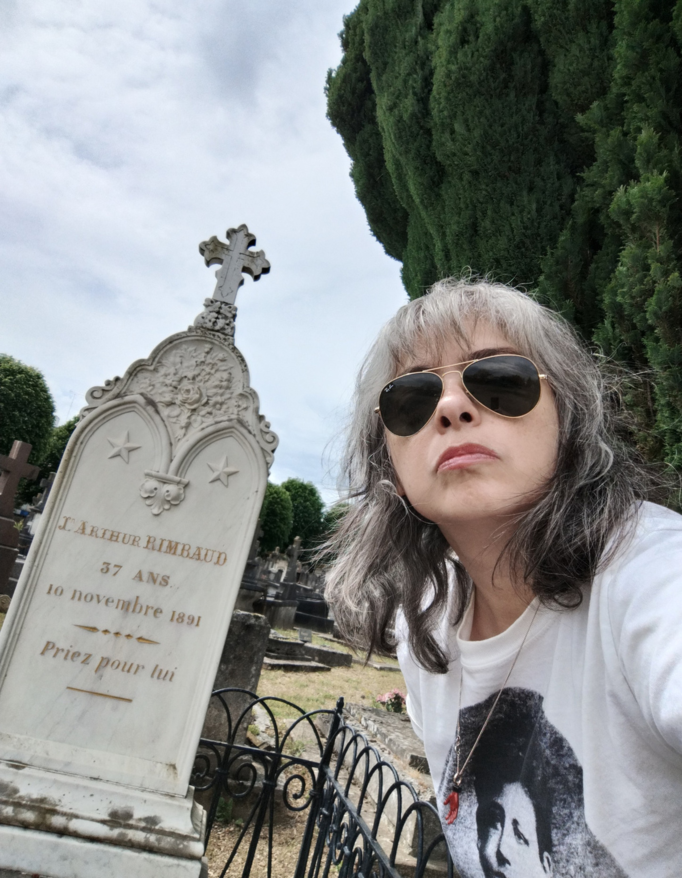 A selfie photo of a woman with shoulder length grey and black hair wearing glasses with a headstone in a cemetery