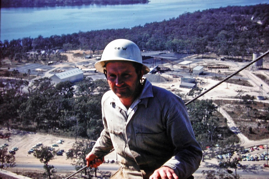 A smiling man on top of a very high cooling tower in a coastal location.