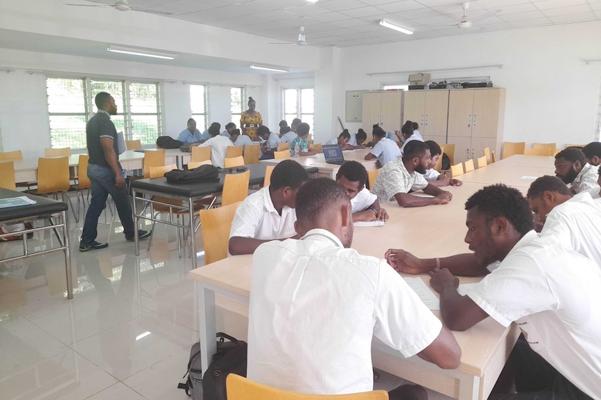 27 people sitting in three groups at desks writing on paper forms in a large, open sunny room.