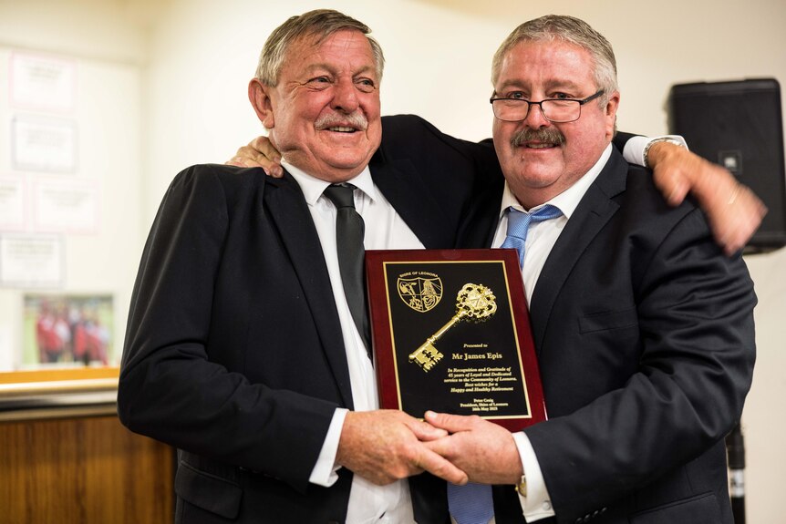Two men embracing with arms around each other showing off a plaque given as a gift to retiring worker.  