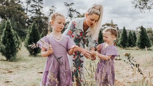 Jo Mcneill bends down picking flowers with her two daughters, wearing purple dresses and holding roses.