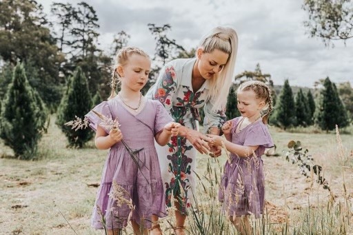 Jo Mcneill bends down picking flowers with her two daughters, wearing purple dresses and holding roses.