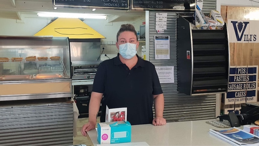 A woman wearing a blue mask is standing behind a deli counter.