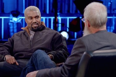 Kanye and David sit facing each other on a stage. The picture is taken from behind David's head so we can only see Kanye