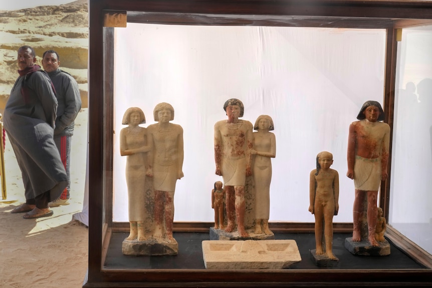 Egyptian statues stand inside a glass case as two men stand off to the side. 