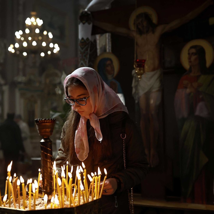 young Ukranian woman wearing headscarf looks down at candles in church setting with painting of Jesus crucifixion in background