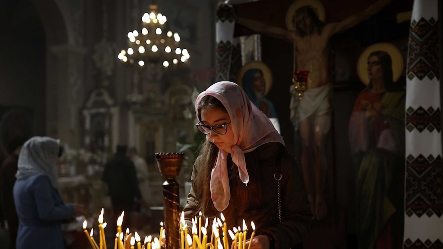 young Ukranian woman wearing headscarf looks down at candles in church setting with painting of Jesus crucifixion in background