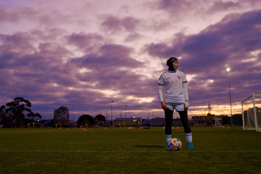 Adiba stands on a Melbourne soccer field in the evening with a ball at her feet wearing a white uniform