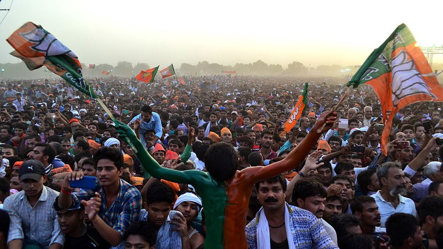 Indian man painted in green and orange holds two flags in front of huge crowd