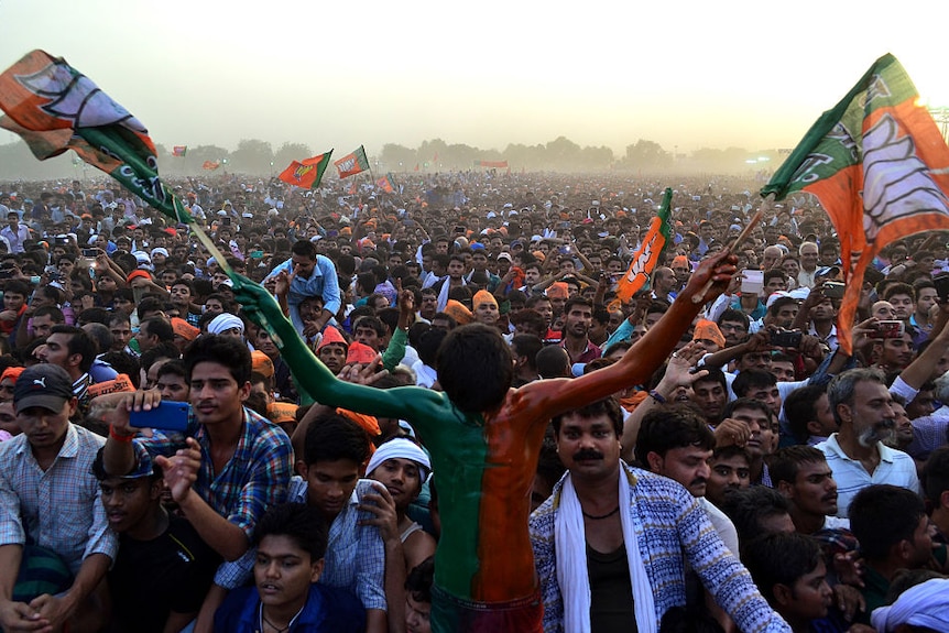 Indian man painted in green and orange holds two flags in front of huge crowd