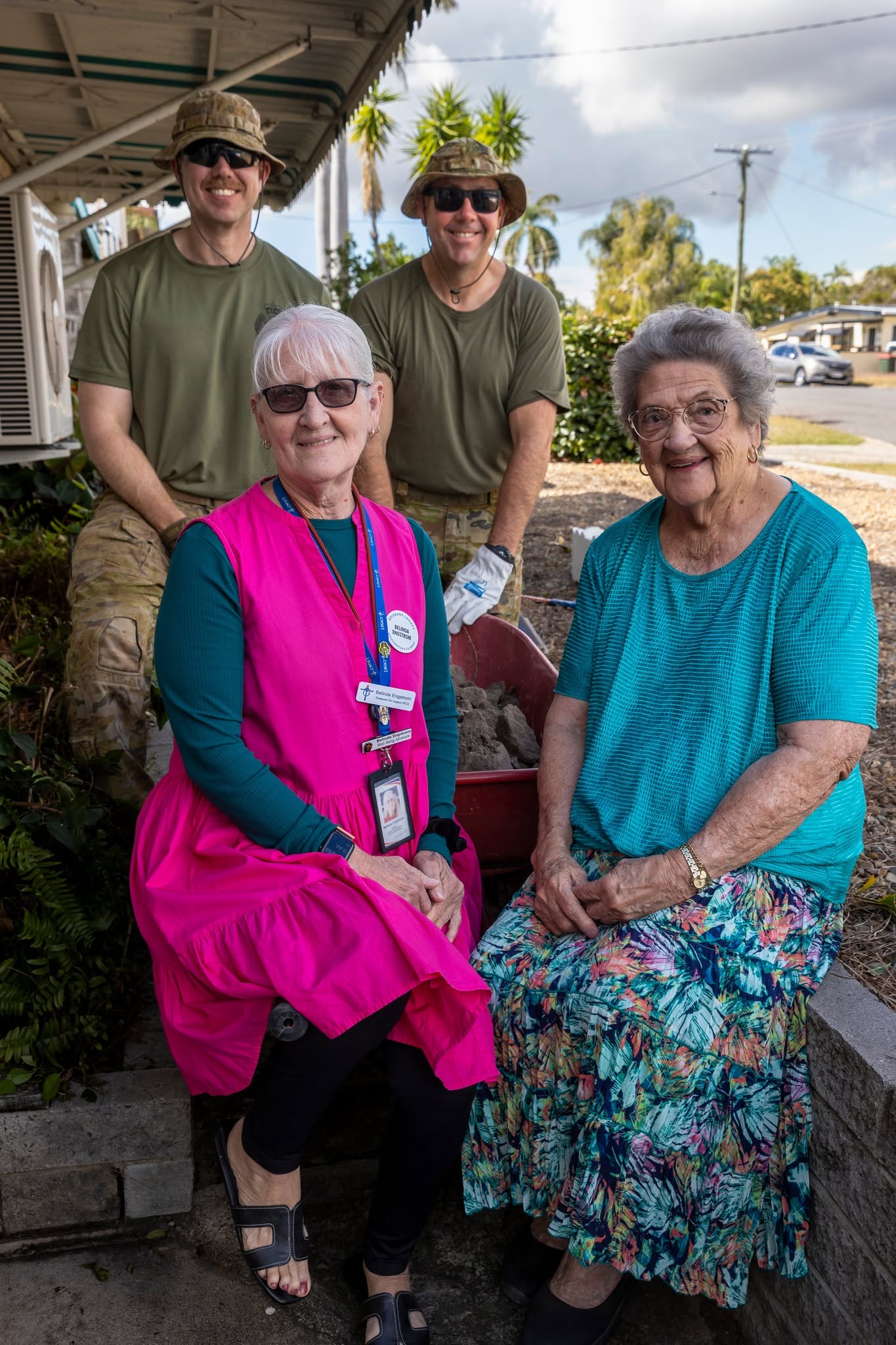 In a front garden, two older woman sit and smile at the camera in front of two standing soldiers in fatigues.