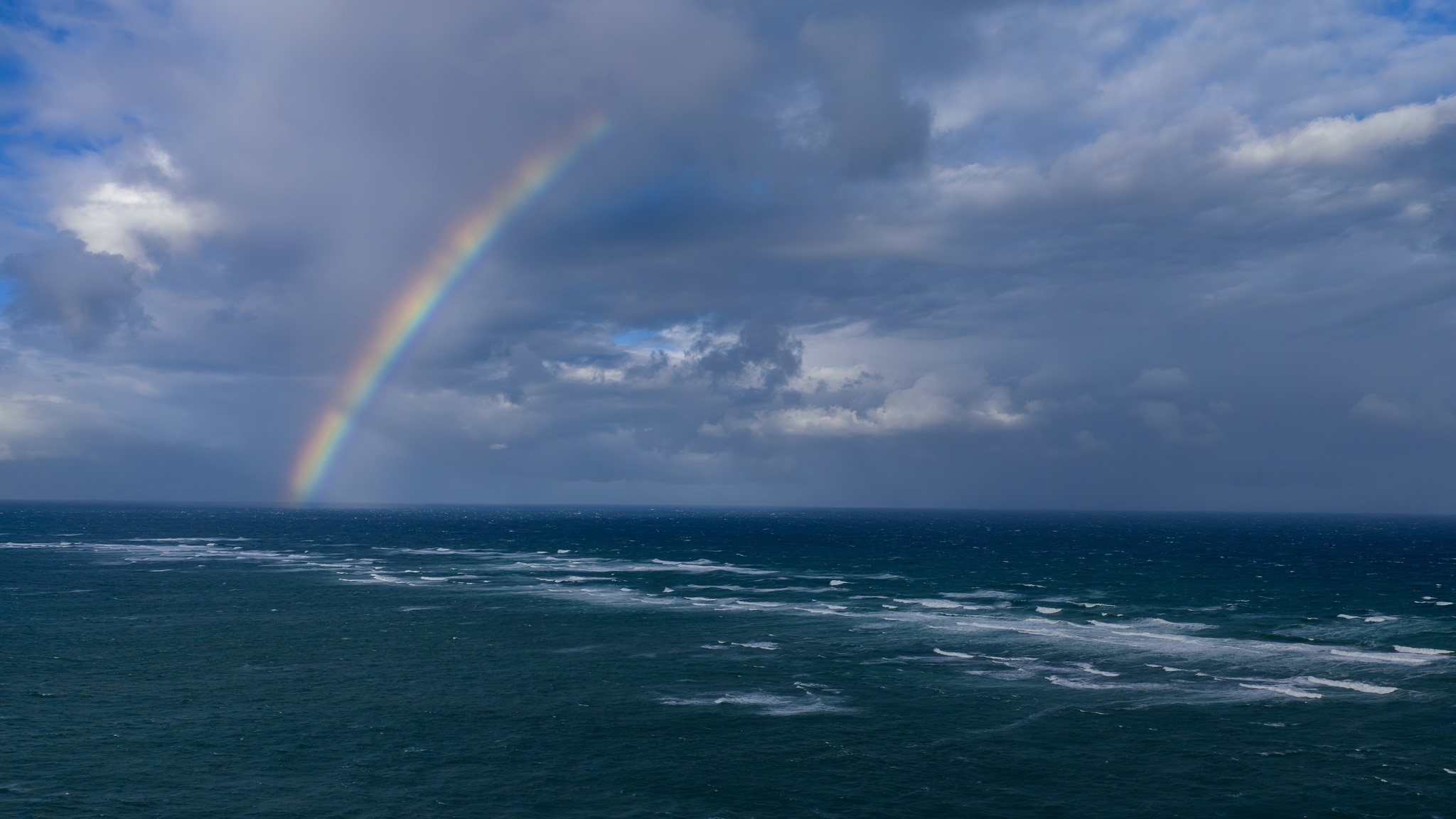 Image of a rainbow in the sky above a stormy ocean with choppy waves. 