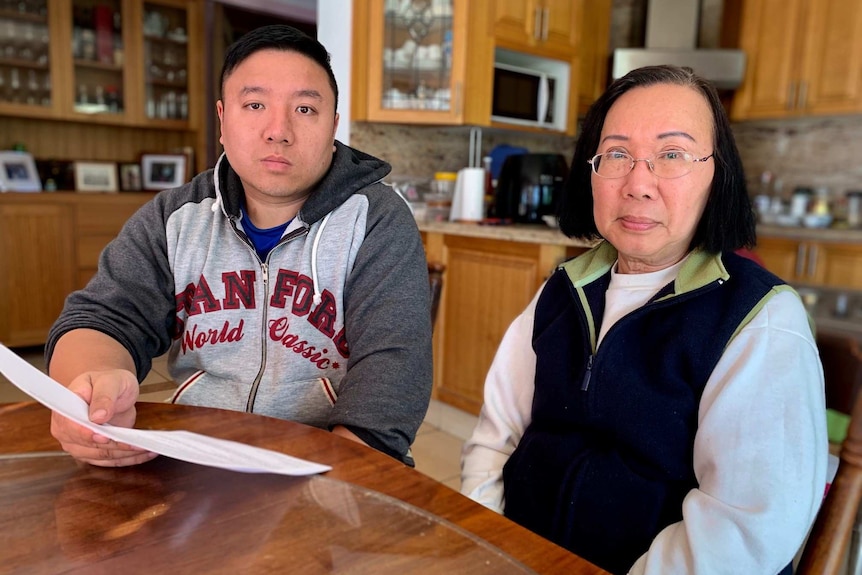 A man in his 30s and woman in her 60s stare ahead at the camera while sitting in their kitchen.