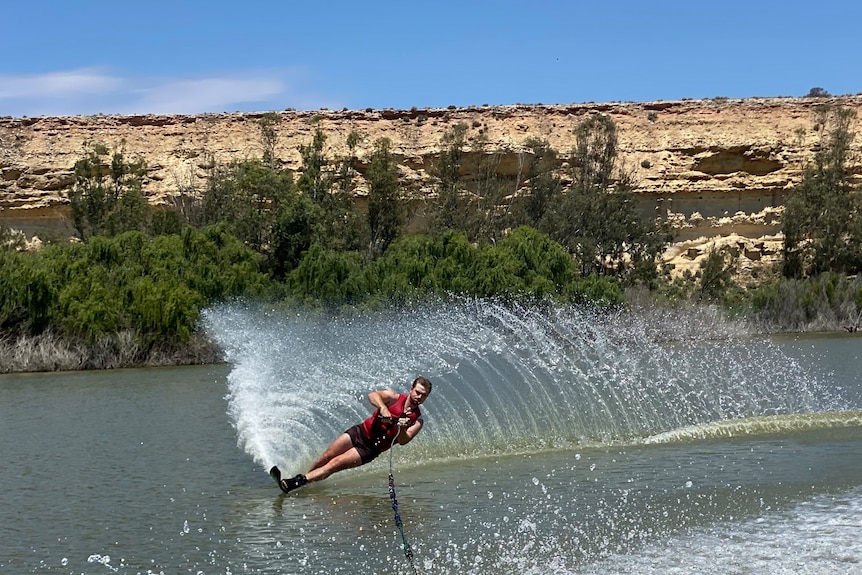 A young man water skiing on the river, with cliffs behind him.