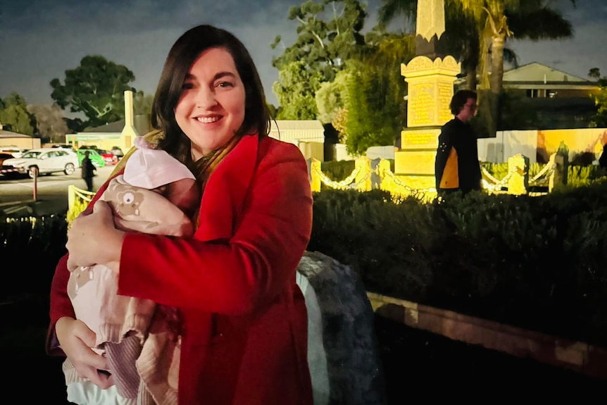 A female politician holding a baby in a carrier stands in front of a war memorial