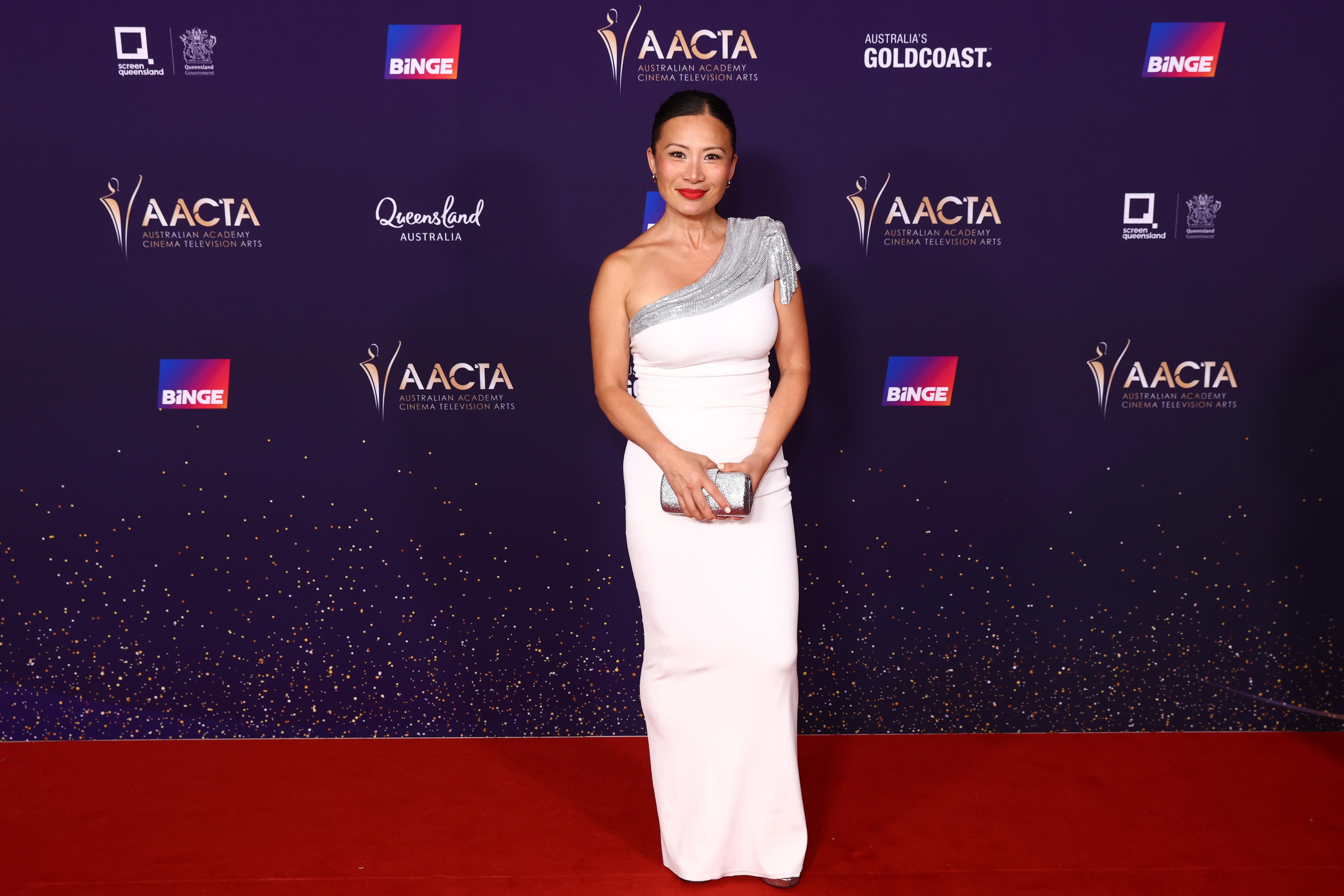 Poh Ling Yeow in a white long dress with a silver drape over her shoulder, a silver purse and red lipstick, hair tied back
