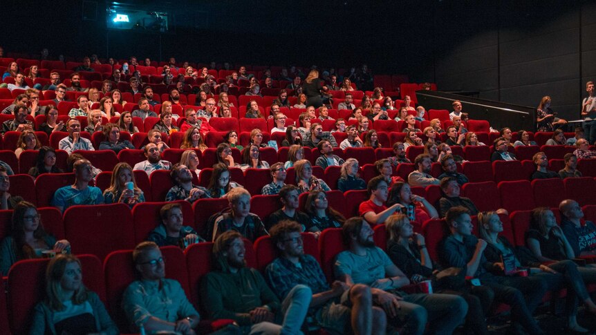 Dozens of people seated in the red chairs of a cinema watching a movie.