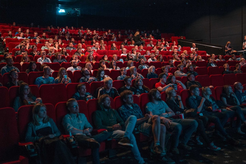 Dozens of people seated in the red chairs of a cinema watching a movie.