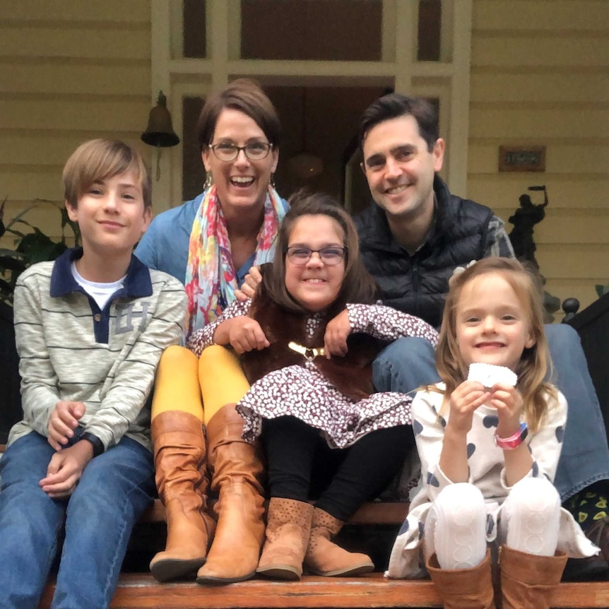 A family of two parents and three children sit on the front steps of a house, smiling at the camera.