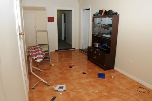 Room with an ironing board on the left wall, a display cabinet on the right and debris, including an iron, strewn on the floor