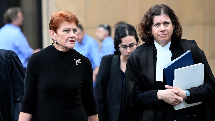 An older woman with red hair wearing all black walks alongside a barrister, carrying paperwork, outside a busy courtroom.