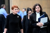 An older woman with red hair wearing all black walks alongside a barrister, carrying paperwork, outside a busy courtroom.