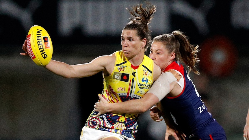 An Adelaide AFLW player holds the ball away in one hand as she is tackled around the waist by a Melbourne player. 