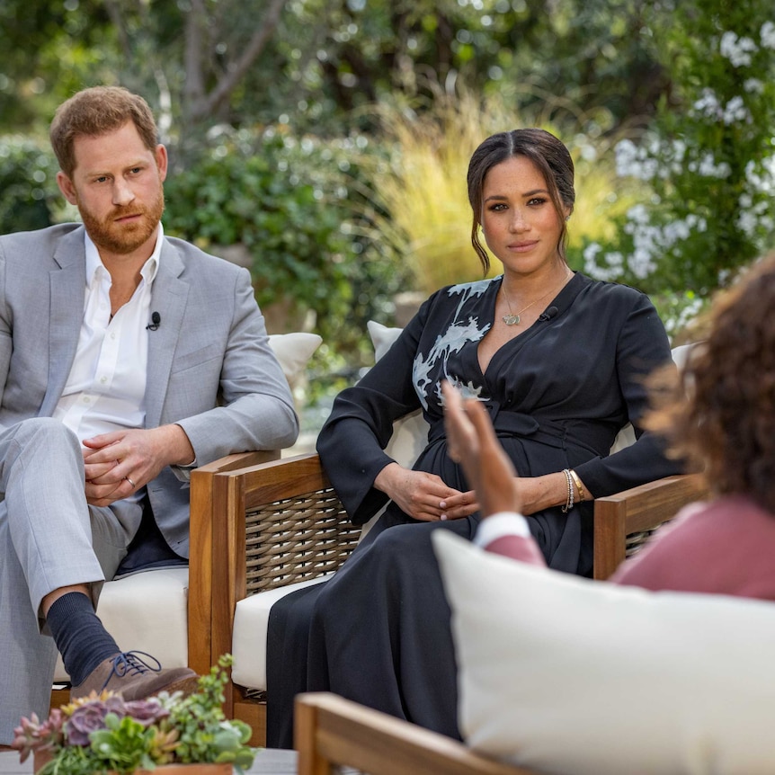 Prince Harry and Meghan Markle sitting in chairs in a garden facing Oprah Winfrey