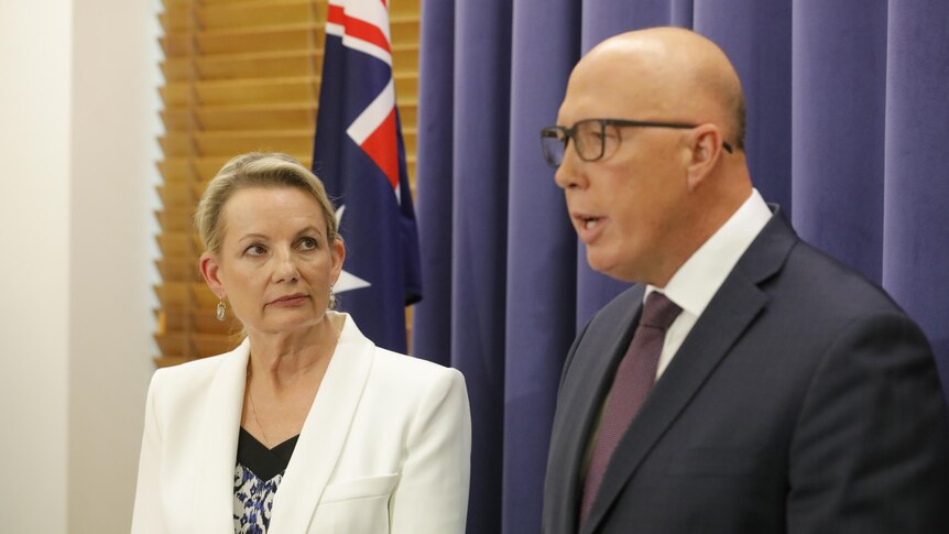 Sussan Ley stands next to Peter Dutton as he speaks in front of a blue curtain, an Australian flag is behind them