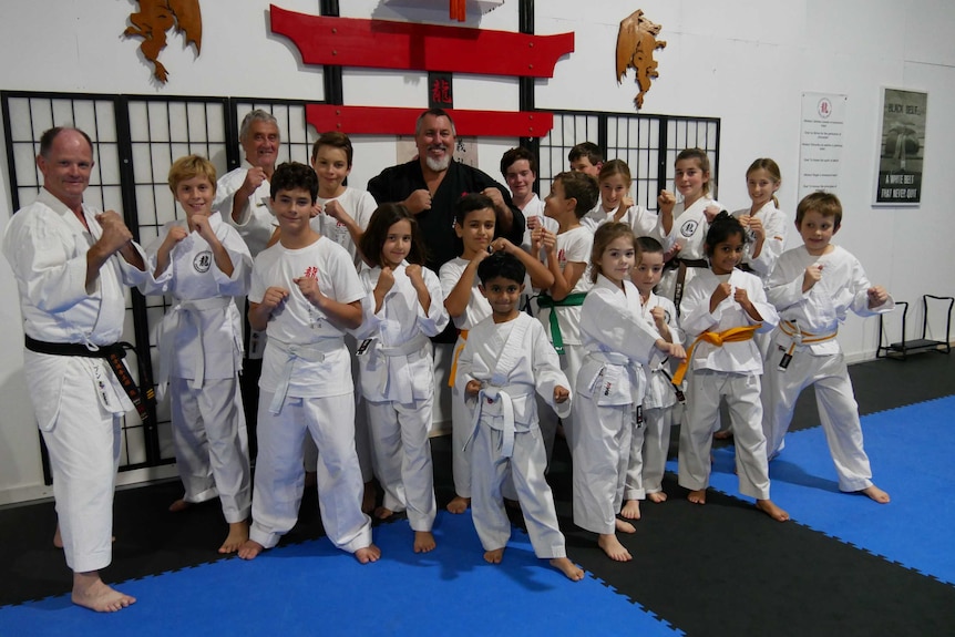 A group of children in white martial arts uniforms standing together smiling with their arms and fists raised.