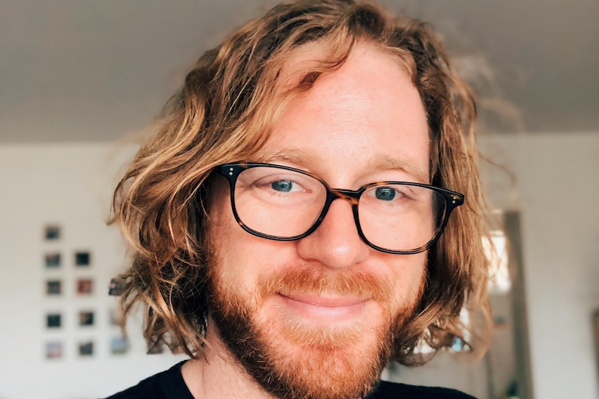 A man with long reddish hair and glasses poses for a portrait.