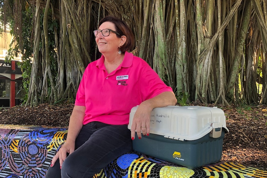 A woman in a pink 'Mission Australia' shirt leans on a first aid box while sitting under a large tree.