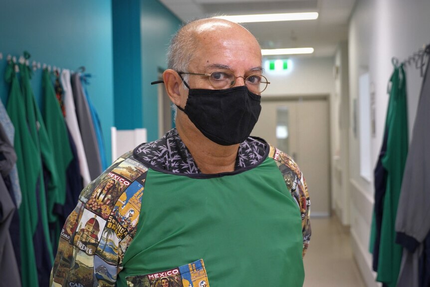 A doctor in a patterned shirt stands in protective gear and a mask, looking at the camera in hospital ward.