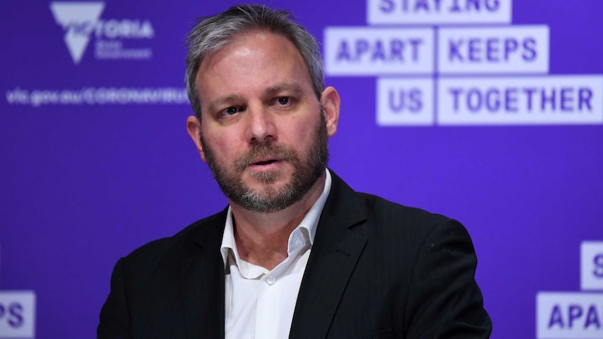 Brett Sutton stands in front of a purple wall with the words 'Staying apart keeps us together' written on it.