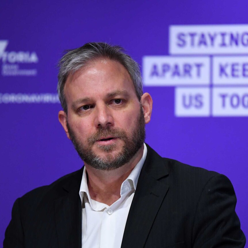 Brett Sutton stands in front of a purple wall with the words 'Staying apart keeps us together' written on it.