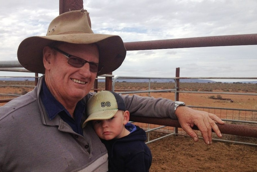 man wearing cowboy has hugs small boy who is his grandson with a sheep paddock in background 