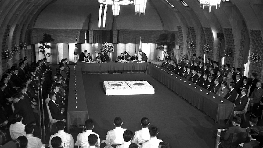 An group of officials assemble in a room draped with flags of Korea and Japan as documents are signed.