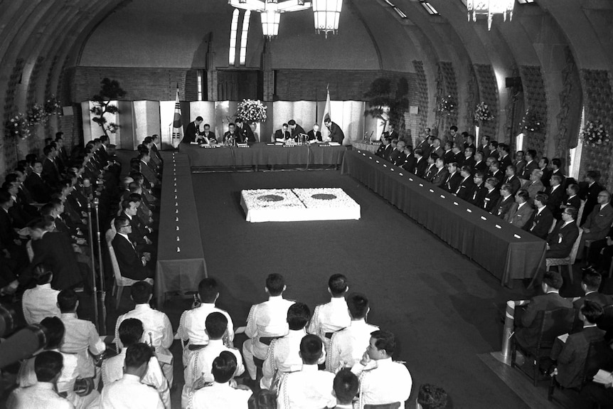 An group of officials assemble in a room draped with flags of Korea and Japan as documents are signed.