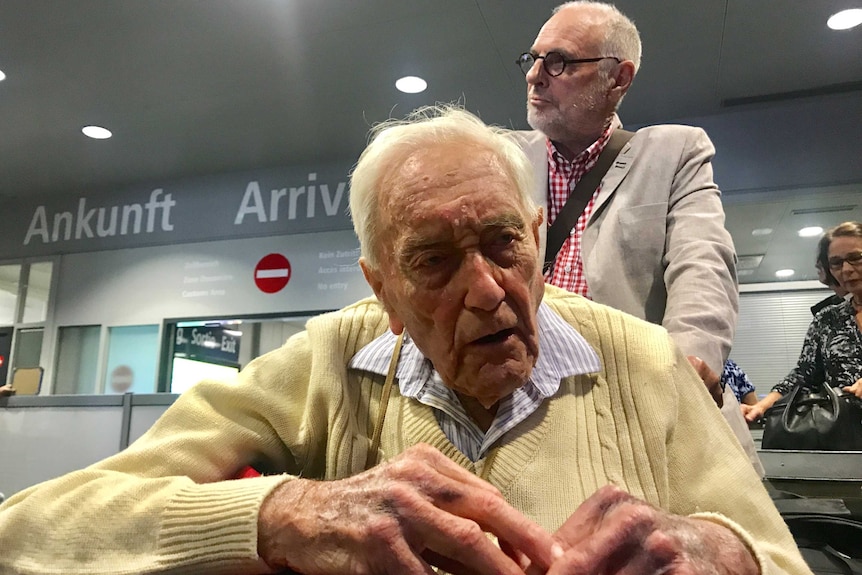 An elderly man in a tan jumper sits on a wheelchair being pushed through an airport by a man in a white coat and black glasses.