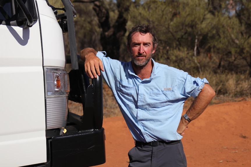 A man in a blue shirt leans against a white truck on a red sand road.