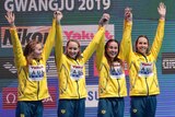 Four female swimmers stand on a medal podium and raises their arms together to celebrate winning gold.