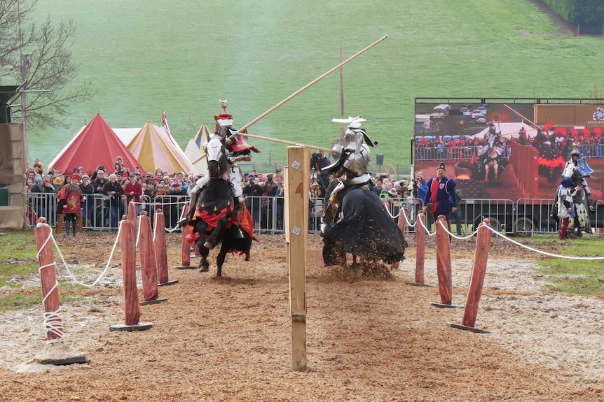 People dressed in armour on horseback joust in front of a crowd and a green lawn.