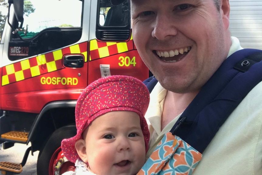Selfie of Nathan baby-wearing his smiling daughter Georgina, with a fire engine in the background.